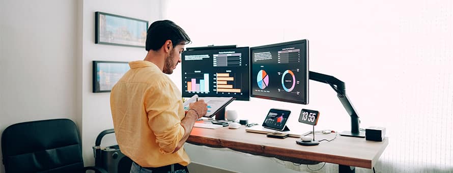 A man in an orange shirt standing at a desk looking at two monitors and researching how to become an economist