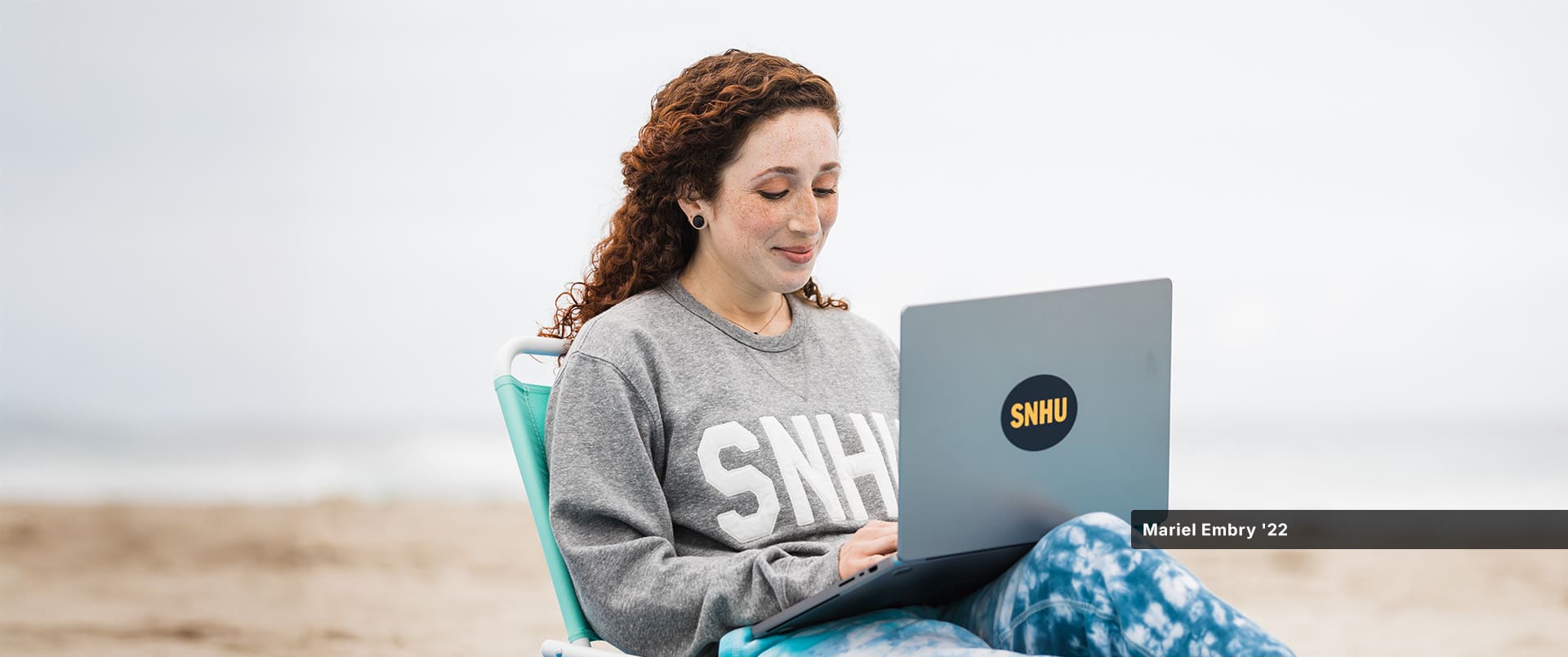 Mariel Embry, who earned her degree in creative writing and English from SNHU, wearing a grey SNHU sweatshirt and writing on her laptop while sitting on the beach.