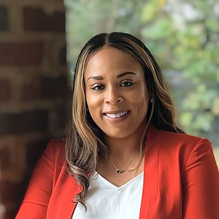 Jolivette Wallace, Associate Dean of Business at SNHU, wearing a red blazer and leaning against a brick column.