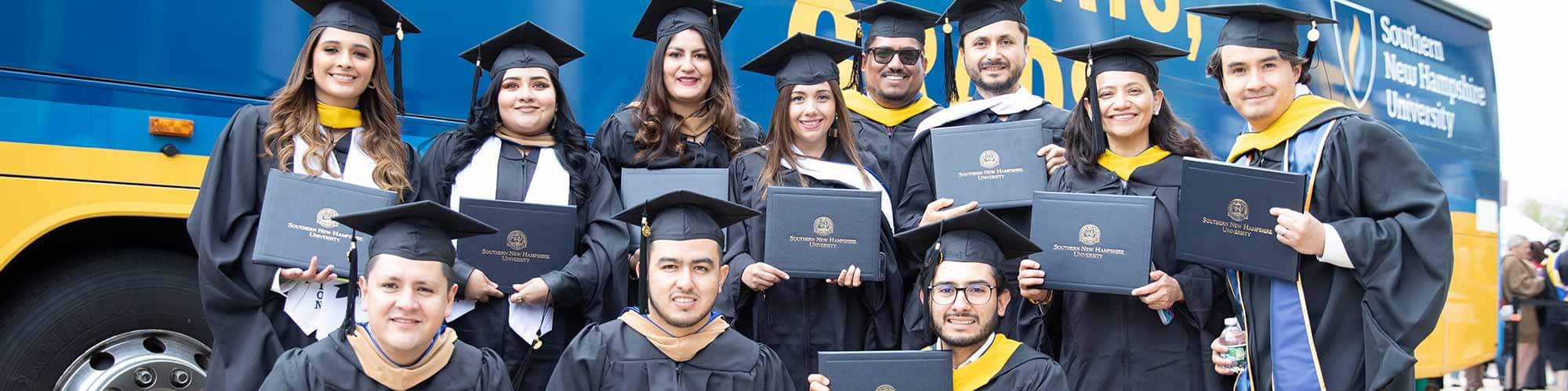 Eleven SNHU grads in caps and gowns holding their diplomas up in front of an SNHU branded bus