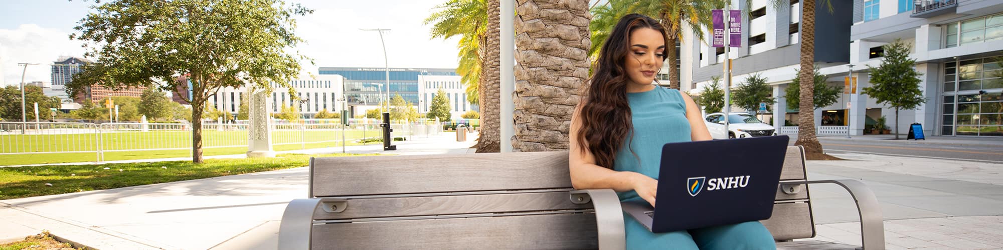 SNHU student and Florida resident Deonna Purrazzo, sitting on a bench in front of a palm tree typing on her laptop.