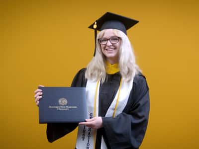 Gigi Bell, who earned her online bachelor's in business administration from SNHU in 2025, wearing her cap and gown and holding her diploma in front of a yellow backdrop.
