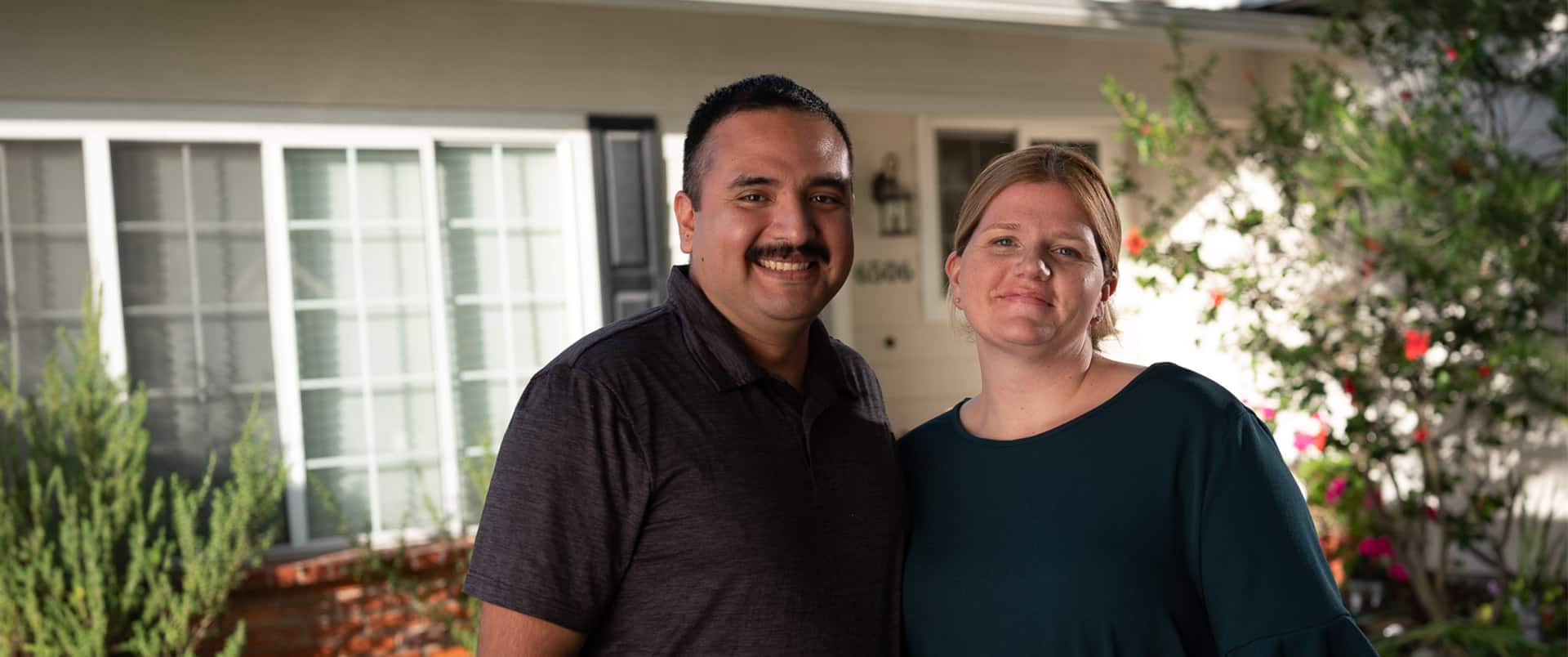 Shelly & Salvador Villa, a married couple who earned their degrees from SNHU in 2019, standing next to one another in their front yard with plants and a white house in the background.