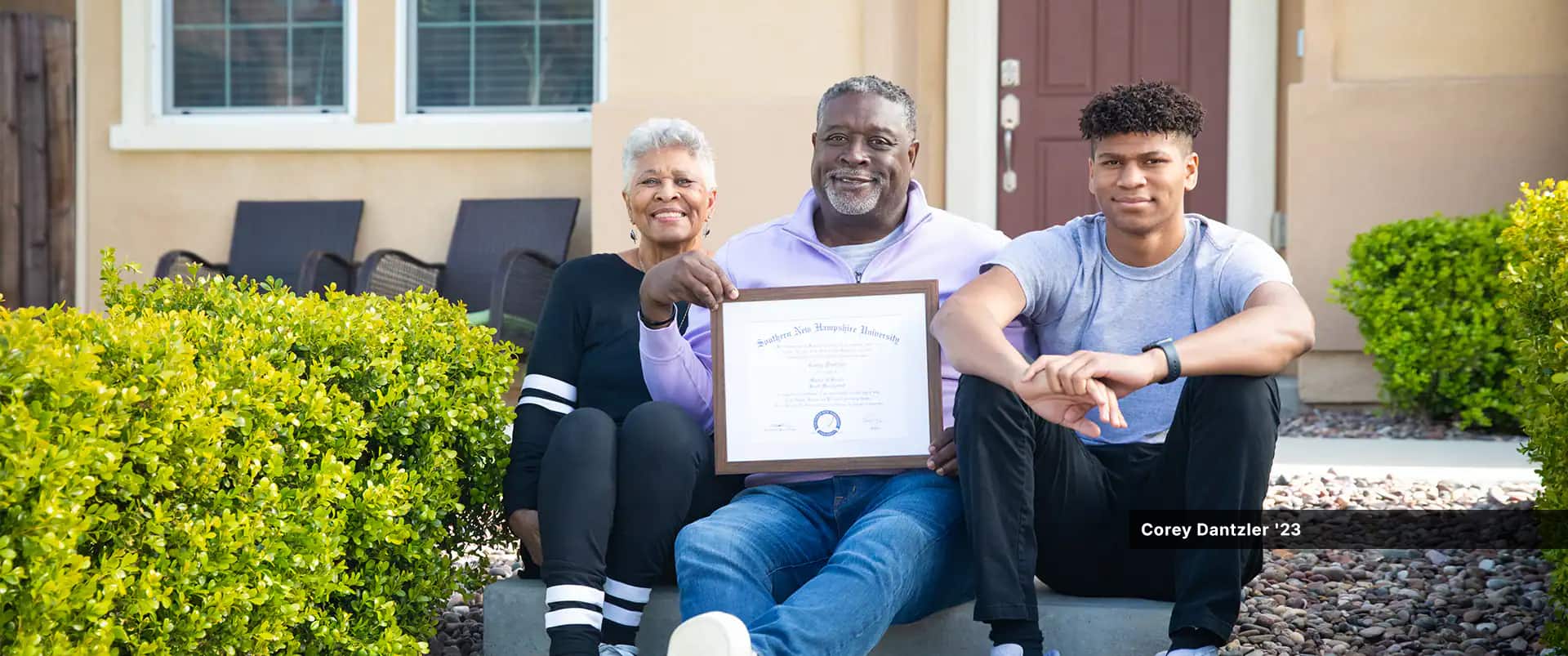 Corey Dantzler, who earned a graduate certificate in athletic administration in 2023, holding his framed SNHU diploma sitting on stone steps in front of his home with a younger man to his left and an older woman with white hair to his right.