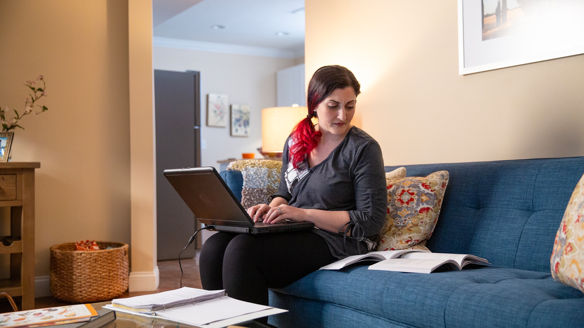 Zepure Kurumlian, who earned her degree from SNHU, sitting on a blue couch in her living room typing on her laptop and looking at an open book on the couch next to her.