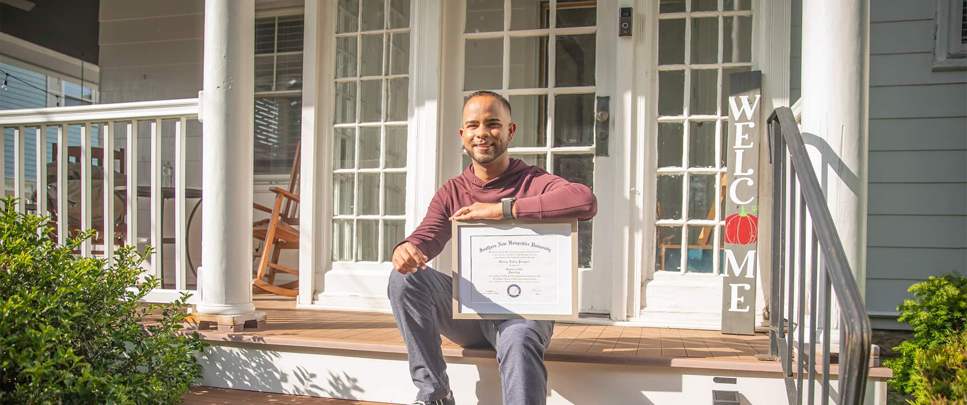 Geremy Laboy, who earned his degree from SNHU, sitting on a porch step holding his framed diploma on his knee with a vertical welcome sign in the background.