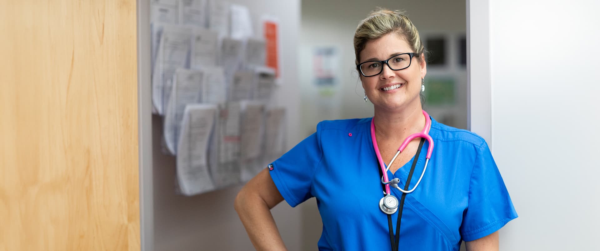 Kristina Libby, who earned her degree from SNHU, standing in a medical office wearing blue scrubs and glasses with a bright pink stethoscope around her neck.