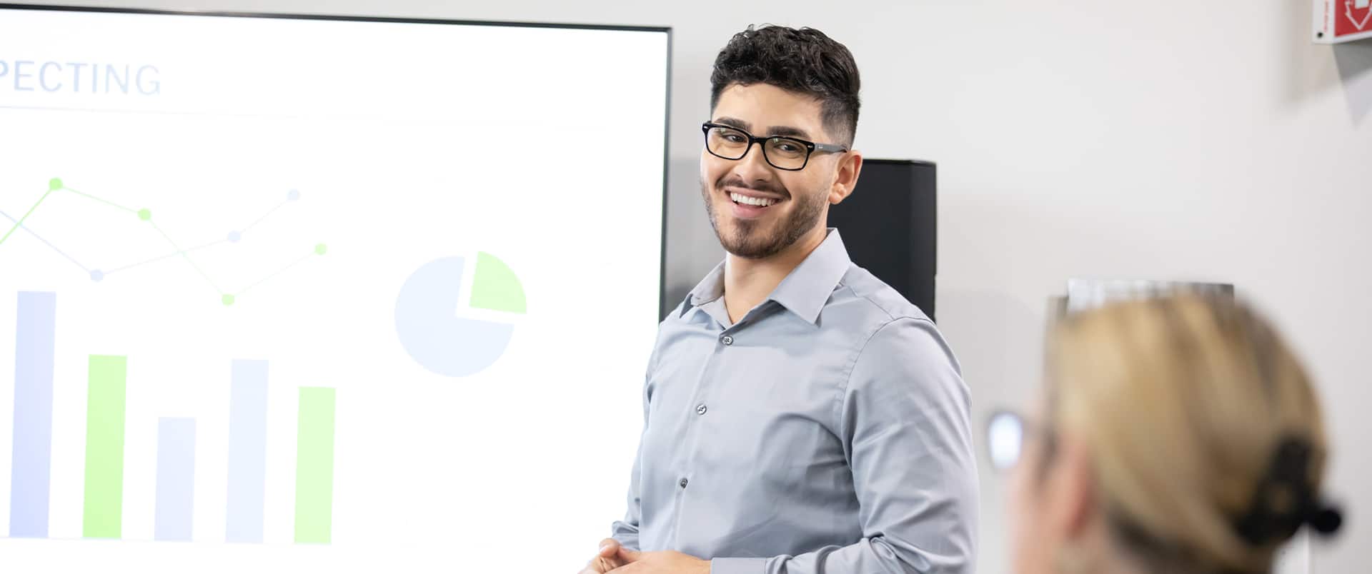 Naeem Jaraysi, an SNHU alum, wearing a blue buttondown shirt and standing in front of a projector screen displaying several charts and graphs.
