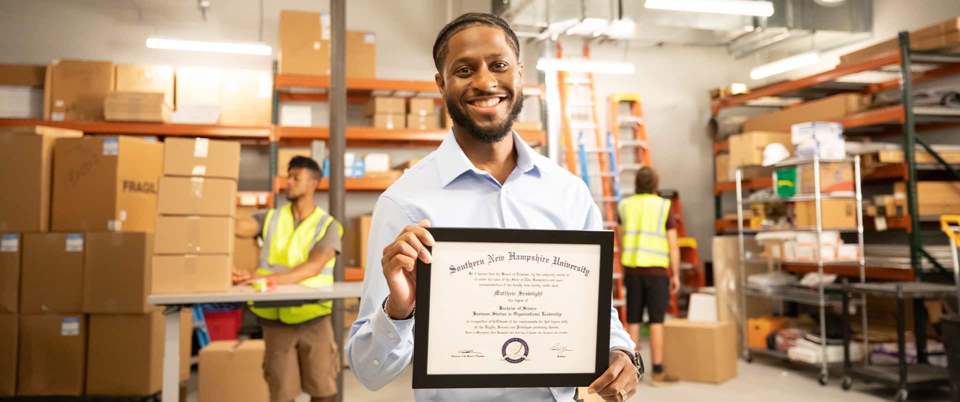 Matt Seawright, who earned a degree in operations and supply chain management in 2019, holding in a warehouse holding his framed SNHU degree with two warehouse workers in the background.
