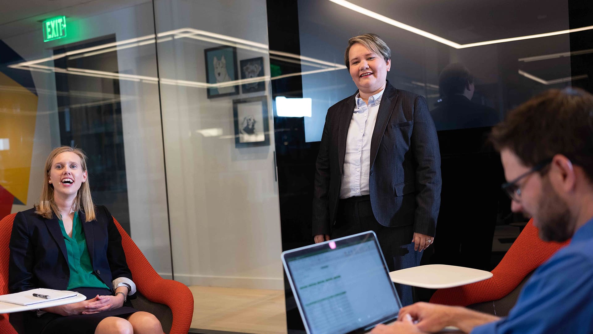 Alaine Garcia, who earned her degree in 2020, wearing a dark blazer with a laughing woman seated to her right and a man in the foreground working on a laptop