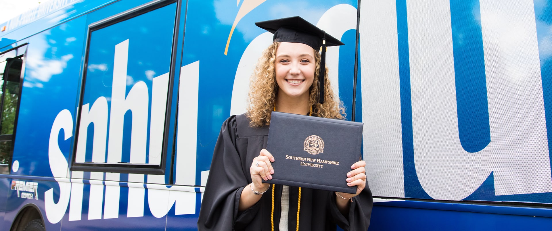 Joanne Coffey, wearing her cap and gown and holding her diploma while standing in front of the SNHU-branded bus.