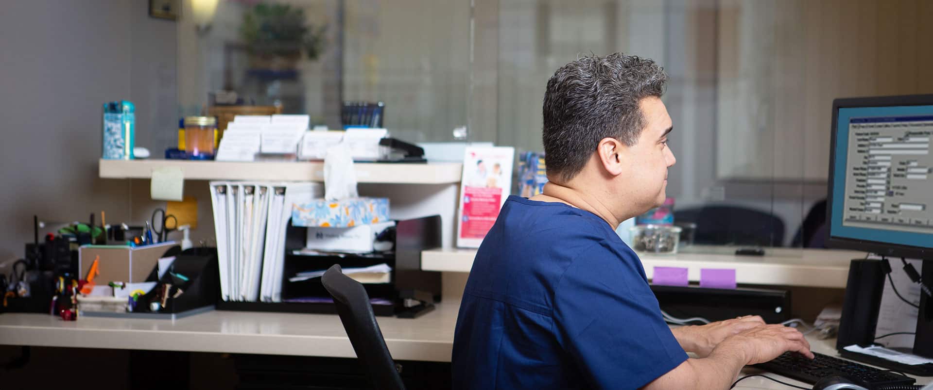 A man wearing medical scrubs typing on a desktop computer with medical files and pamphlets in the background.