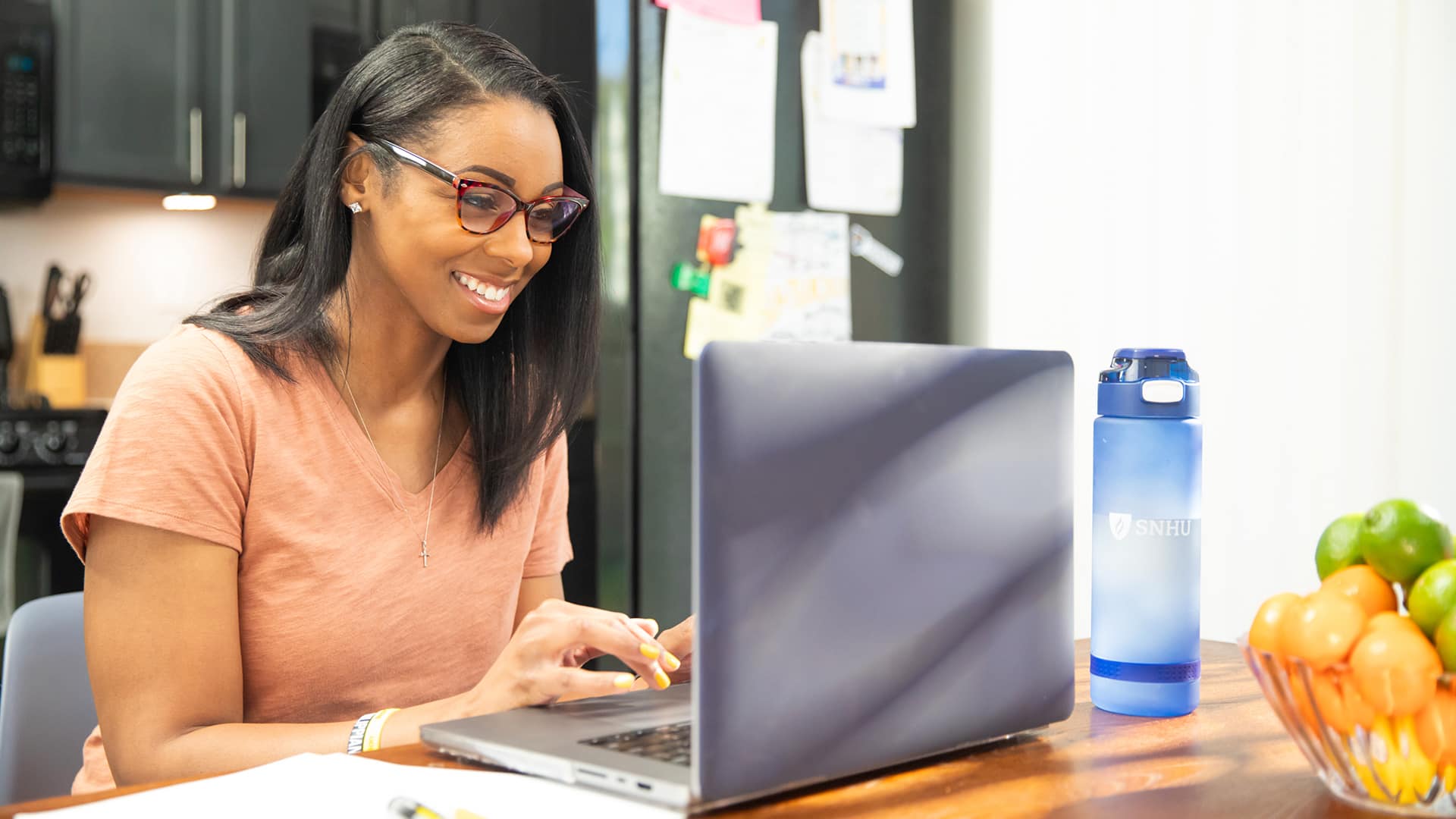 A woman working on a laptop on her kitchen counter with blue SNHU water bottle next to her.