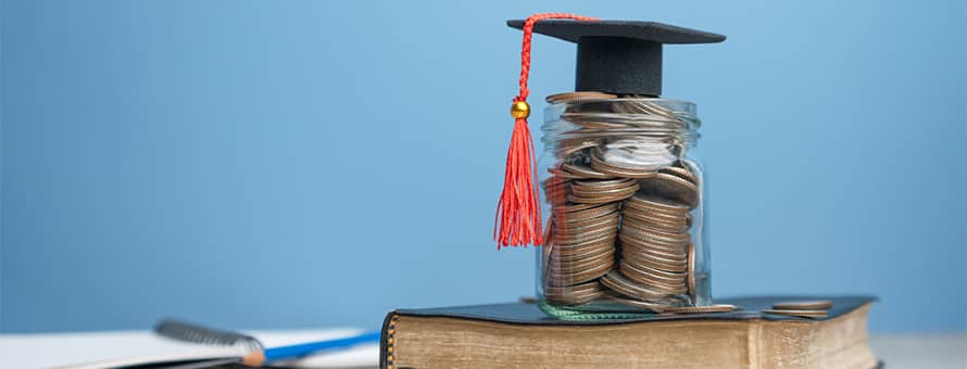A book beneath a stack of coins with a tiny graduate cap on top.