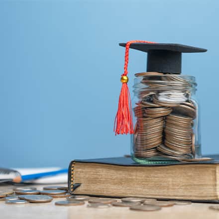 A book beneath a stack of coins with a tiny graduate cap on top.