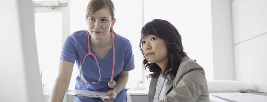 A woman who became a licensed practical nurse, wearing a stethoscope, holding a clipboard and leaning toward a computer screen a healthcare professional is using.