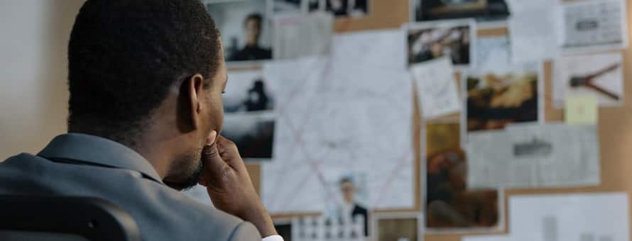 A detective sitting in a chair looking at a bulletin board going over evidence 