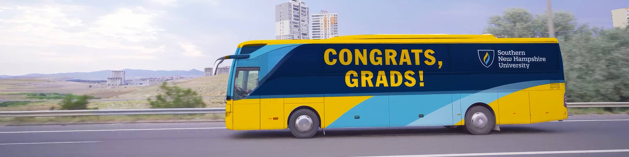 A yellow and blue coach bus with the words Congrats, Grads and Southern New Hampshire University on the side driving down a highway.