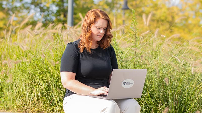 Ali Lamoureux-Rule, who earned her healthcare administration degree from SNHU in 2022, typing on her laptop with a field of tall grass behind her.