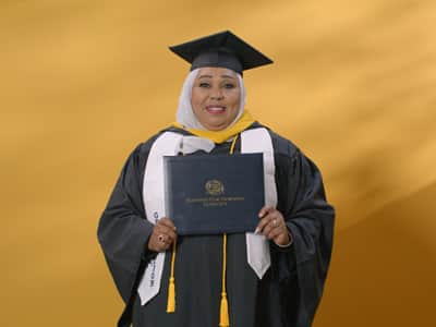 Jamila Aithammousaid, who earned her bachelor's in nursing from SNHU in 2024, wearing her cap and gown and holding her diploma in front of a yellow backdrop.