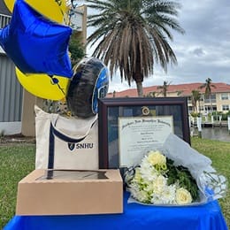 Joan Donovan’s SNHU diploma on a table with flowers, a bag with an SNHU logo and balloons.