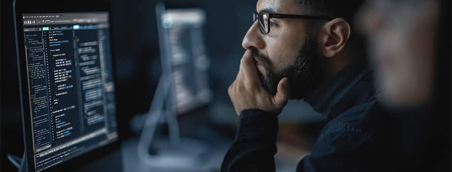 A man wearing glasses looks at code on a computer screen with his chin resting on his hand.