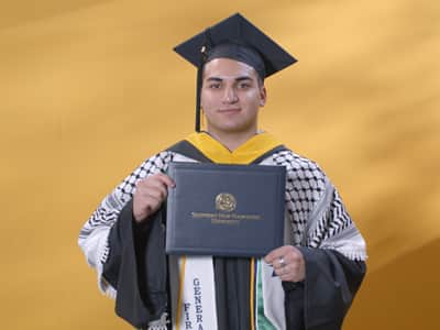 Jonathan Oubenslimane, who earned his bachelor's in business administration from SNHU in 2024, wearing his cap and gown and holding his diploma.