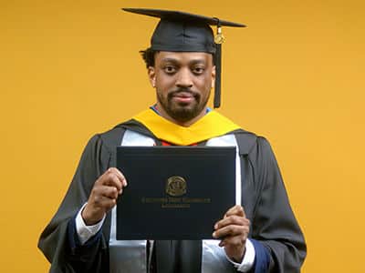 Josiah Lee, who earned his online master's in cybersecurity from SNHU in 2025, wearing his cap and gown and holding his diploma in front of a yellow backdrop.