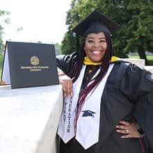 Karen Raquel Quezada, who earned her master's in psychology from SNHU in 2021, wearing her cap and gown and standing next to her diploma.