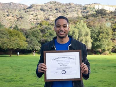 Keyon Tuiteleleapaga, who earned his psychology bachelor's from SNHU in 2022 and graduate degree in 2024, standing in a green field holding his framed diploma.