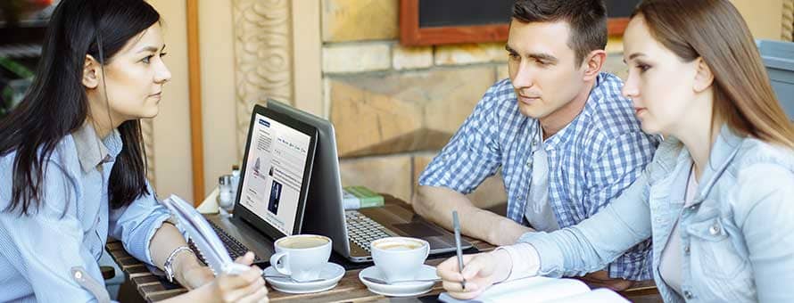 Woman holding up a notebook to two colleagues while her laptop is open next to her