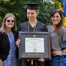 Loretta Gray dressed in her cap and gown, holding her SNHU diploma between two friends.