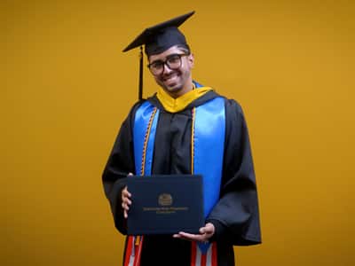 Luis Zayas, who earned his online bachelor's in healthcare administration from SNHU in 2025, wearing his cap and gown and holding his diploma in front of a yellow backdrop.