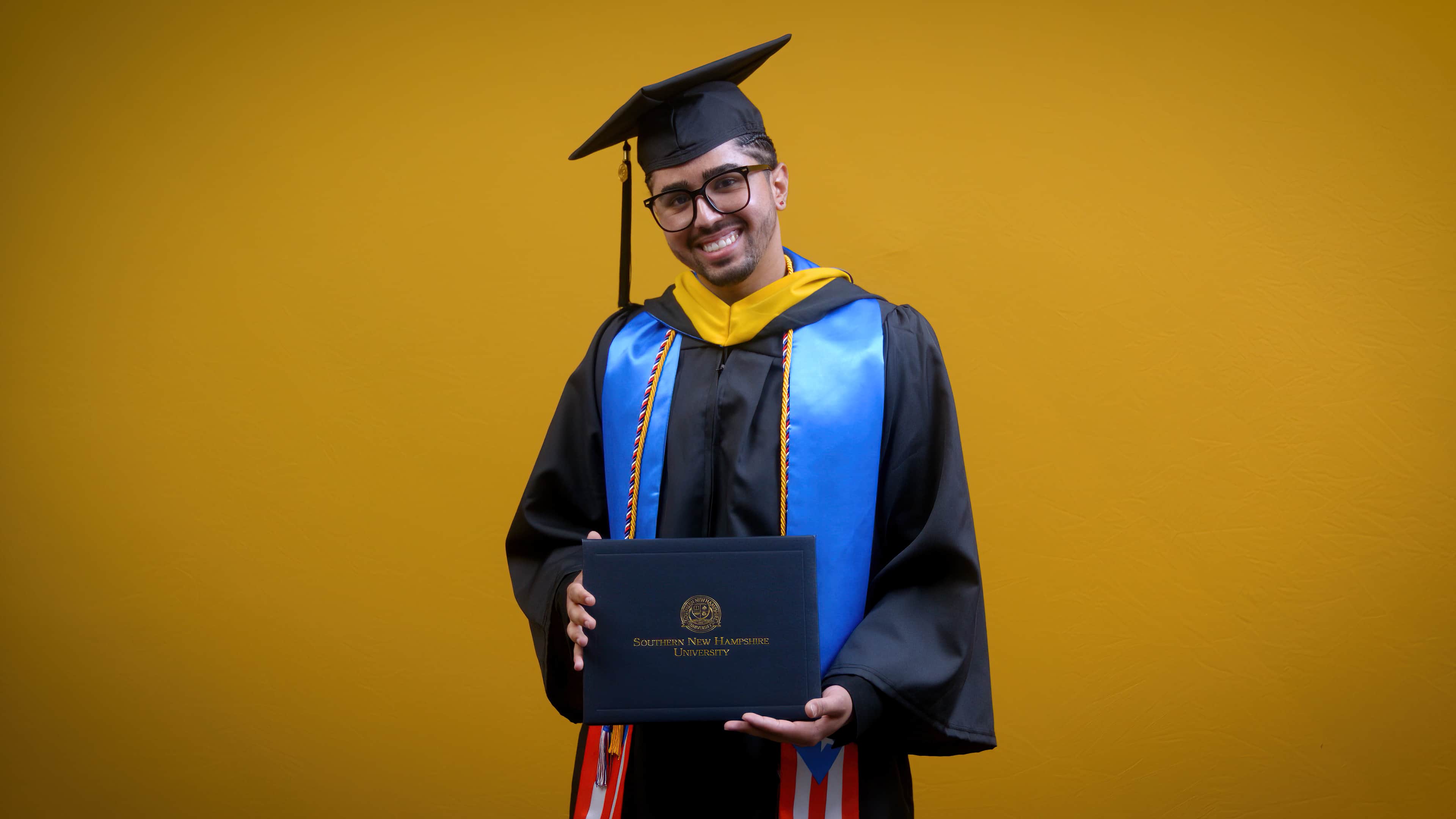 Luis Zayas, who earned his bachelor's in healthcare administration in 2025, wearing his cap and gown and holding his diploma in front of a yellow backdrop.