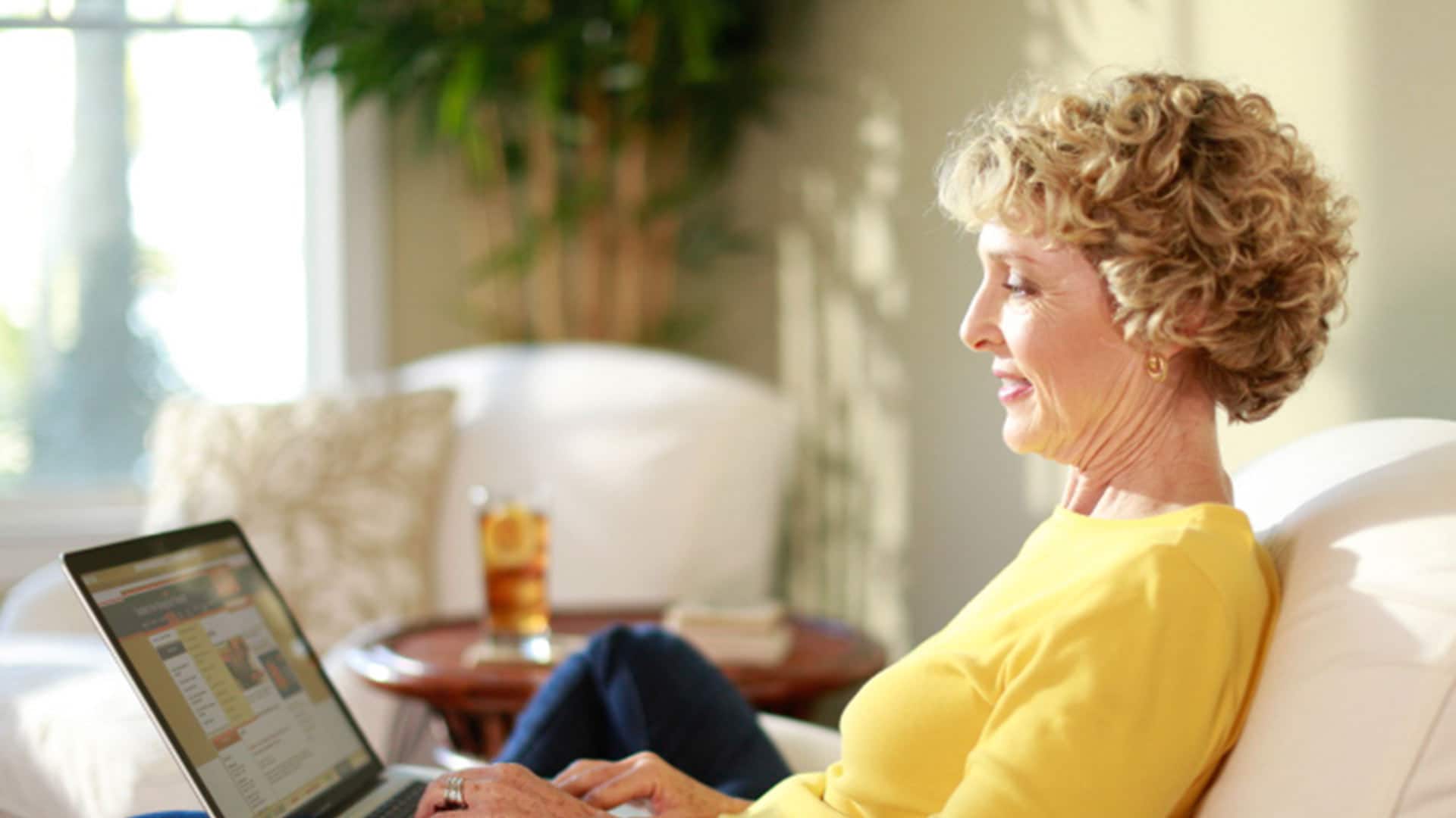 Susan Ryan, who earned her degree from SNHU in 2016, wearing a yellow shirt sitting on a couch typing on her laptop with a glass of iced tea with lemon on the table in the background.
