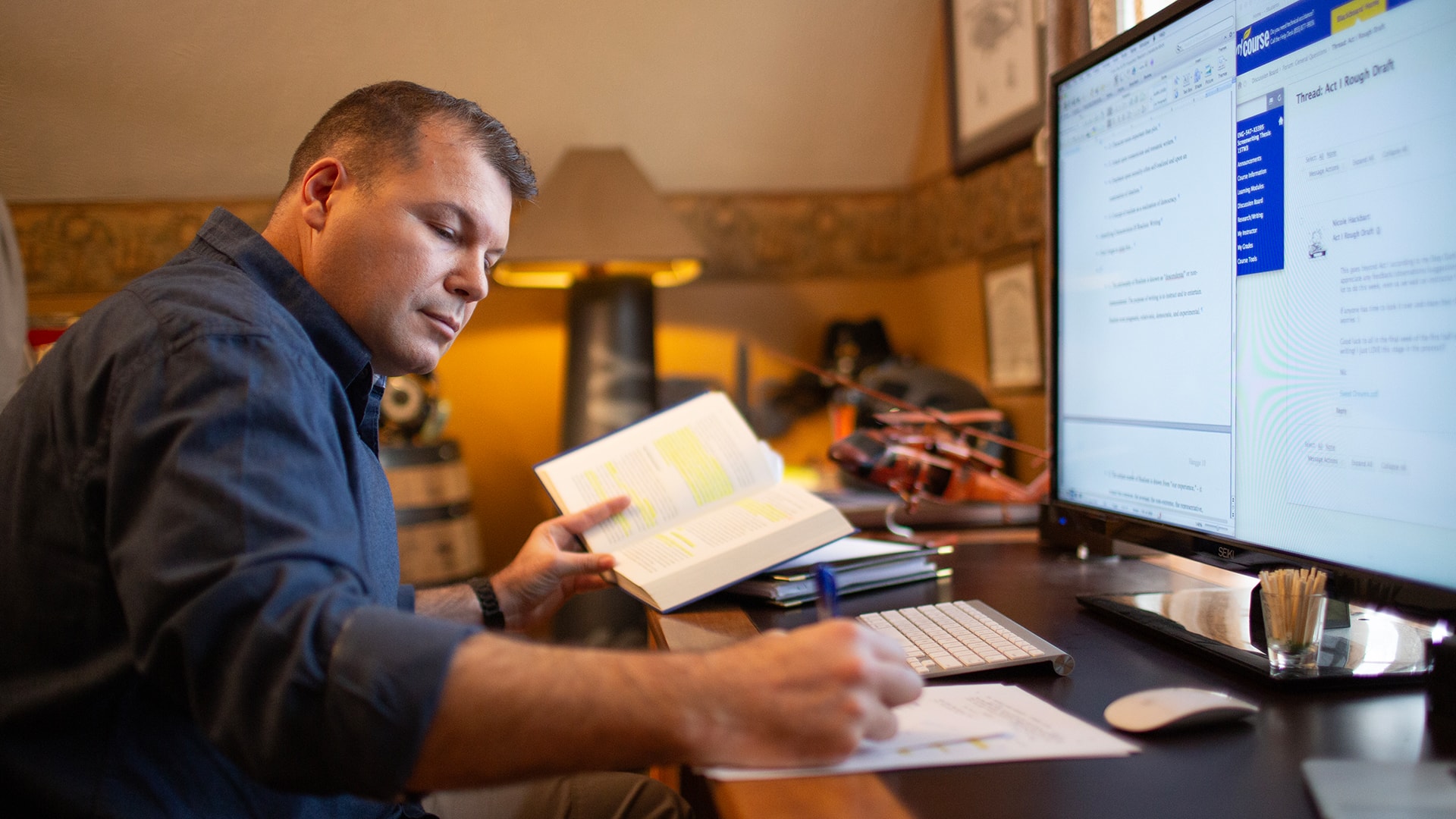 Michael Hangge, who earned his degree from SNHU in 2015, at the desk in his home office in front of a computer monitor, holding open a text book with some passages highlighted and taking notes on a piece of paper.