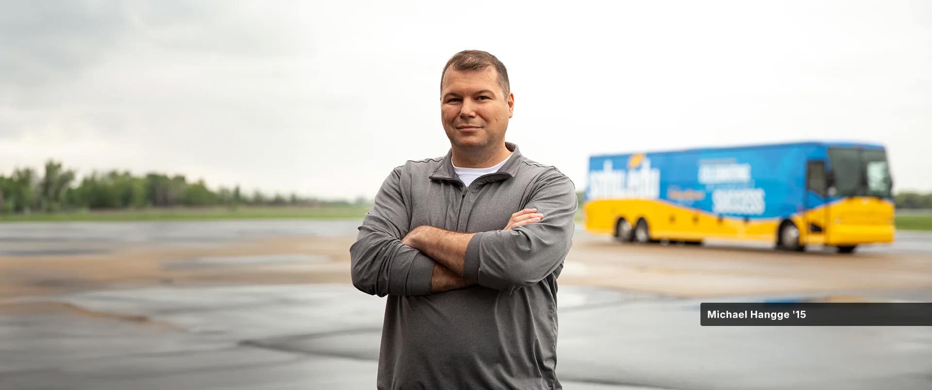 Michael Hangge, who earned his degree from SNHU in 2015, wearing a grey zippered sweatshirt standing with his arms folded with a bus branded with SNHU colors visible in the background.