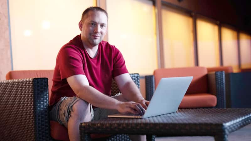 John Roos, who earned his online history degree from SNHU in 2018, wearing shorts and a red T-shirt, sitting in a chair and typing on his laptop.