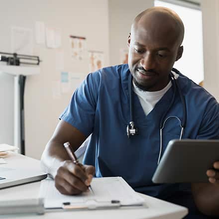A male nurse wearing blue scrubs and a stethoscope writing in a medical chart.