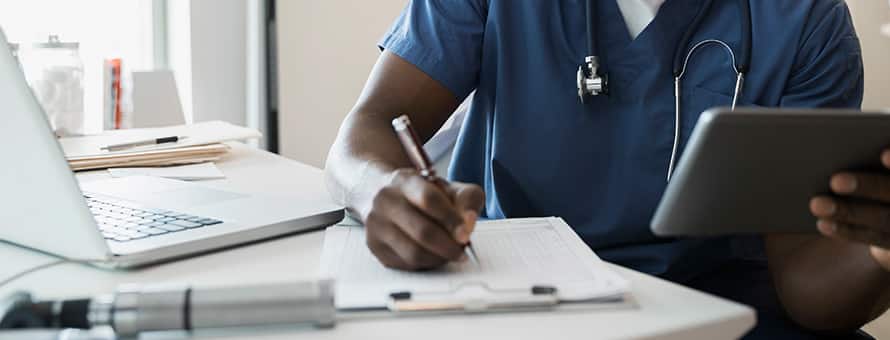 A male nurse wearing blue scrubs and a stethoscope writing in a medical chart.