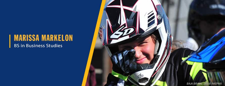 Photo of Marissa Markelon wearing a dirt bike helmet, smiling and pointing toward the camera.