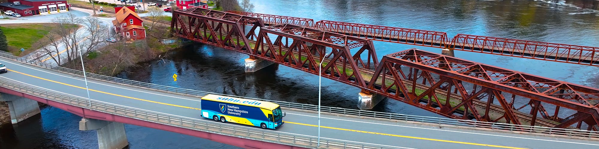 A yellow and blue coach bus with SNHU’s logo on the side seen from above driving over a bridge.