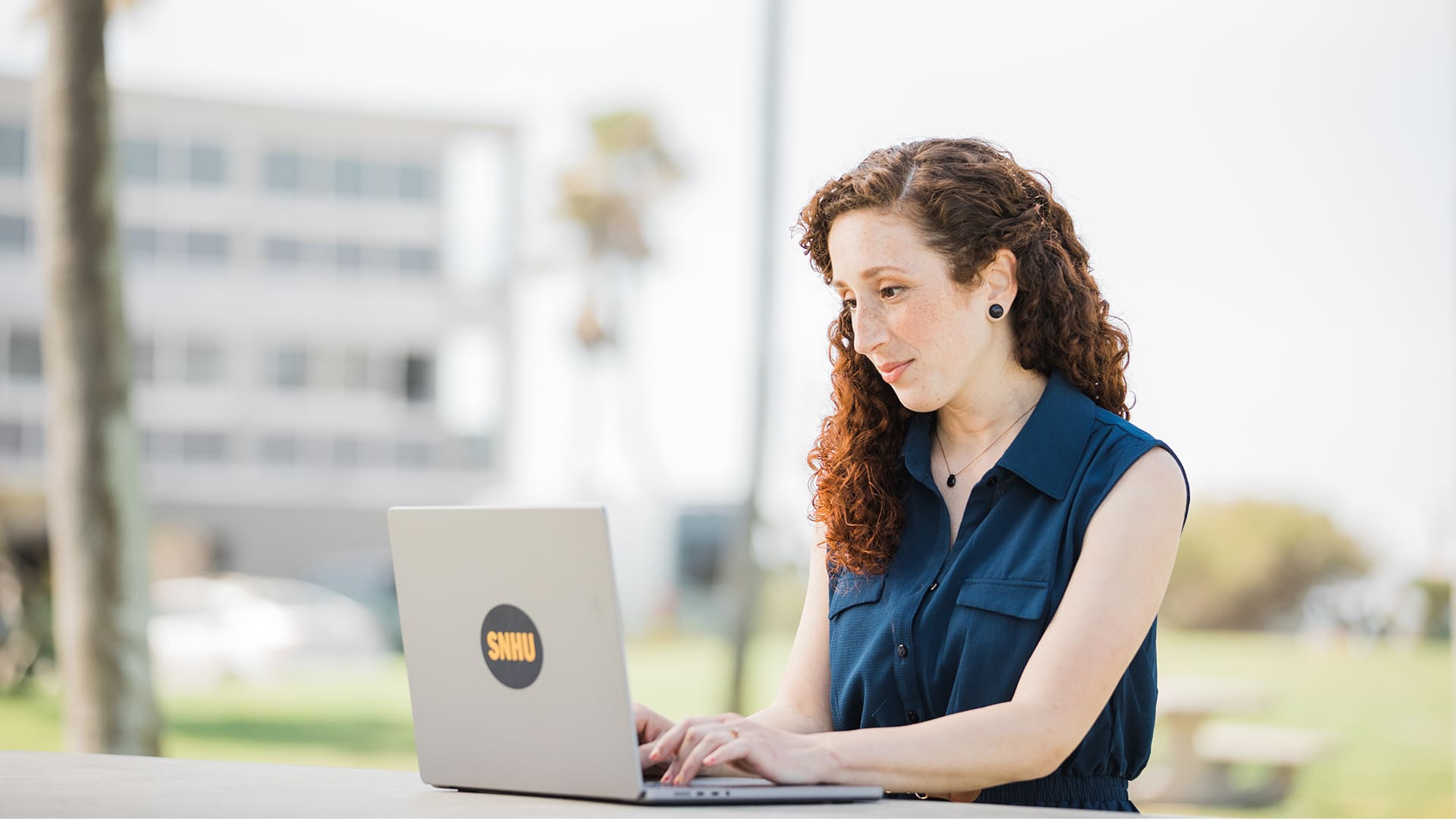 Mariel Embry wearing a navy blue sleeveless shirt working on her laptop with an SNHU sticker on the front .