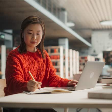 A student in a red sweater using a laptop, pen and notebook to brainstorm what she can do with a master's in English.
