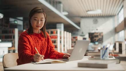 A student in a red sweater using a laptop, pen and notebook to brainstorm what she can do with a master's in English.