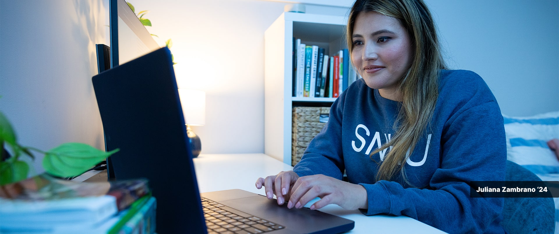 Juliana Zambrano, who earned her degree from SNHU in 2024, wearing an SNHU sweatshirt sitting at a desk typing on a computer with a bookcase in the background.