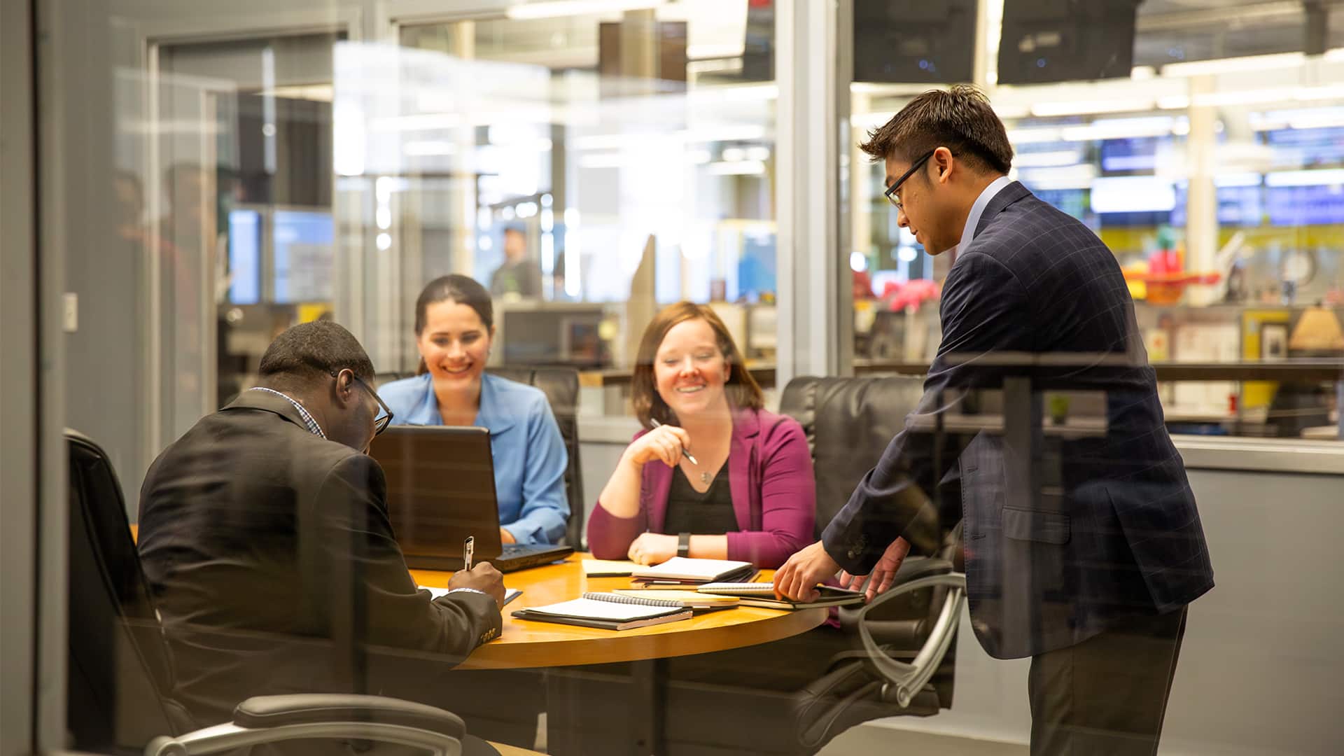 Scott Saktanaset, wearing a dark suit inside a conference room where he is standing and speaking to two women and a man who are seated. 