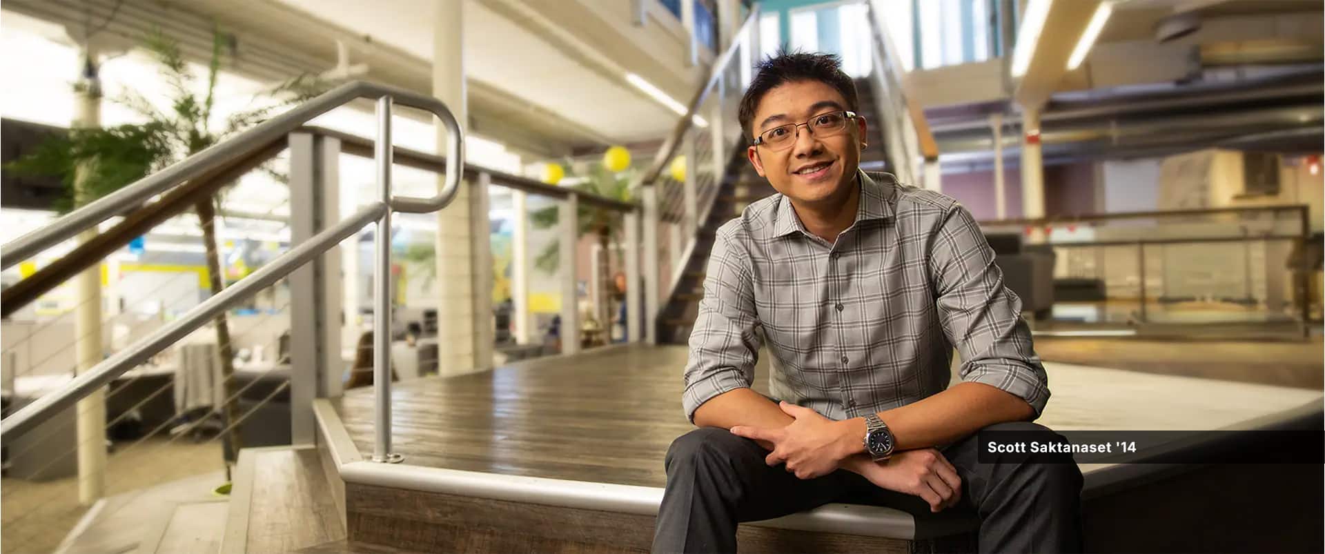 Scott Saktanaset, an SNHU alum, wearing a plaid button-down shirt and wristwatch sitting on a pentagonal platform in the middle of a staircase.