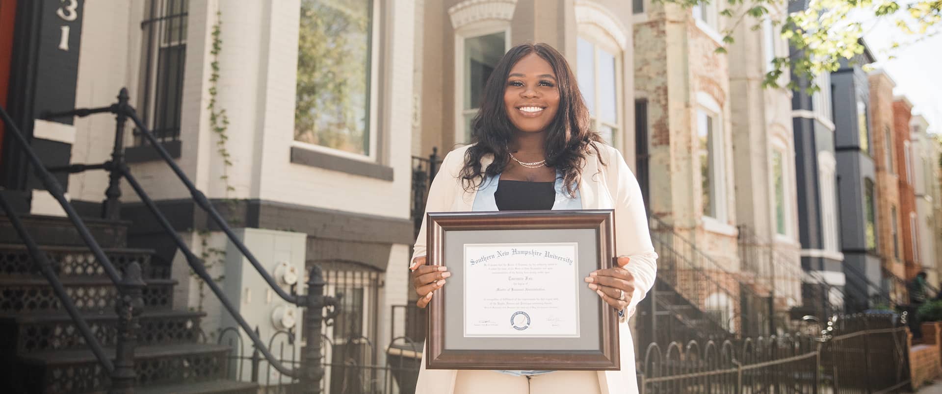Tanzania Fair, who earned her degree in 2020, holding her framed SNHU degree in front of apartment buildings on a city street.