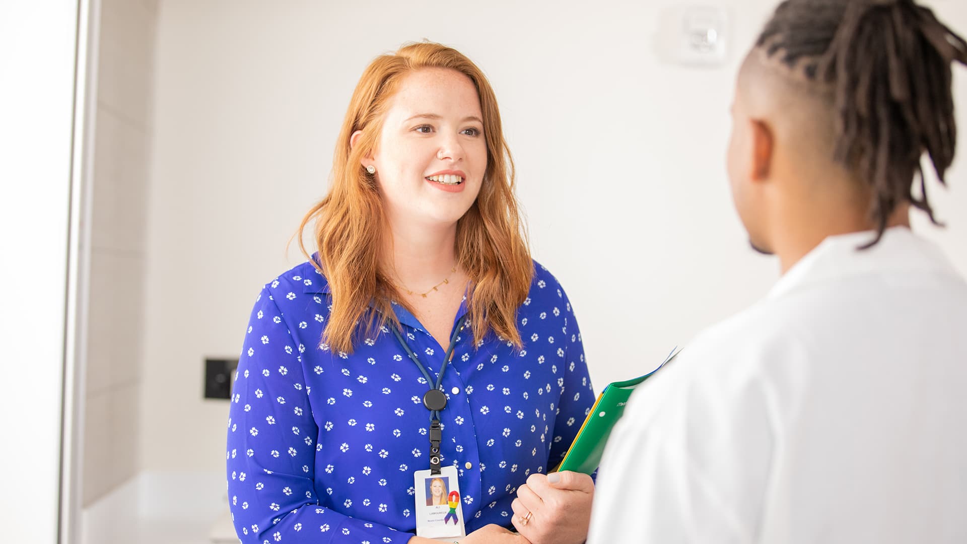 Ali Lamoreux, who earned her degree in 2022, wearing a blue blouse and an ID card on a lanyard around her neck and holding a green folder while talking to a man. 